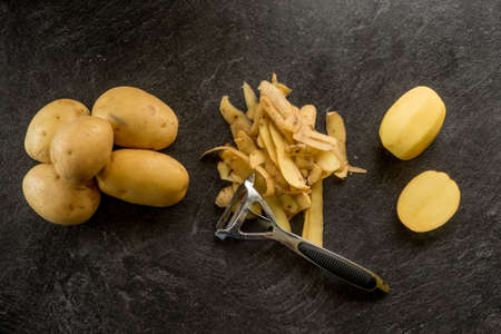 Peeler with peeled potatoes and their skins on textured black background. Photo from aboveの写真素材