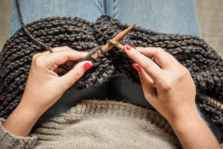 Girl with red nails knitting dark gray wool with wooden needles sitting in a chair, crafts with wool