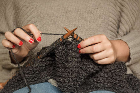 Girl with red nails knitting dark gray wool with wooden needles sitting in a chair, crafts with woolの写真素材