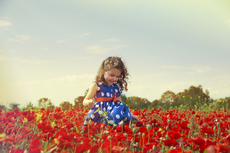 Portrait of adorable smiling brunette girl in blue polka dot dress running in poppy field in sunlightの写真素材
