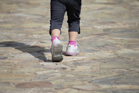 Close-up of unrecognizable little girl in sneakers walking on stone ground in sunlight