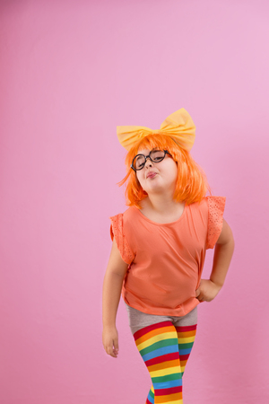 Beautiful seven years girl with an orange wig sticking out her tongue on a pink backgroundの写真素材