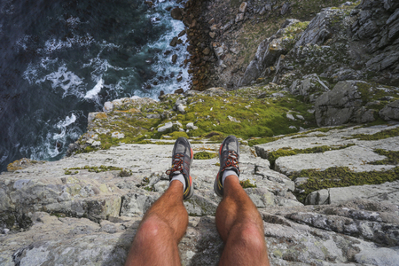 Hiker's legs hanging over the edge of a cliff with the blue ocean far beneath.の写真素材