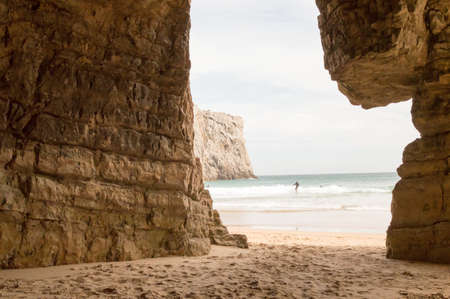 Cave formed on the rocks of Beliche beach in Sagres, Portugal. Photograph taken on June 10, 2016. A surfer is seen walking on the beach with the board and fins.の写真素材