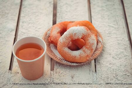 Russian fast food bagel pyshki and paper cup with tea or coffe on wooden table, covered with snow, winter snack in the park.の写真素材