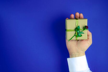 Male hand holding gift box with little red rose. Elegant man in white shirt giving a present wrapped with craft paper and decorated with a flower. Blue background.の写真素材