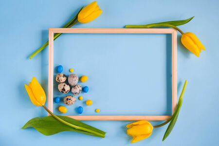 Blue spring and Easter background with a wooden frame, quail eggs colorful candies and yellow tulips. Top view with copy spaceの写真素材