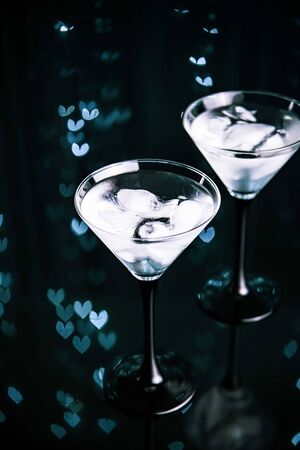 Couple of cocktail glasses with martini drink with ice on trendy black background with heart-shaped bokeh lights. Evening or night date concept.の写真素材