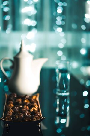 Beautiful bowl in Arabic style full of dry dates fruits, white tea pot and a glass of water, Ramadan concept, on dark blue background and fairy lights.Vertical.の写真素材