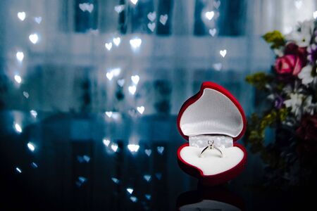 Red heart-shaped velvet box with engagement diamond ring on background with bokeh lights and...