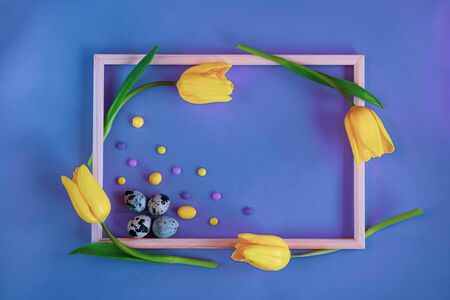 Blue spring and Easter background with a wooden frame, quail eggs colorful candies and yellow tulips Top view with copy spaceの写真素材