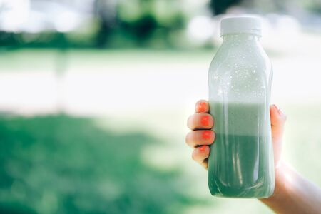 Closeup of womans hand with bright orange manicure holding takeout container of green tea laminaria smoothie with natural background in bright sunny day. Back to normal concept. Takeout cafe.の写真素材