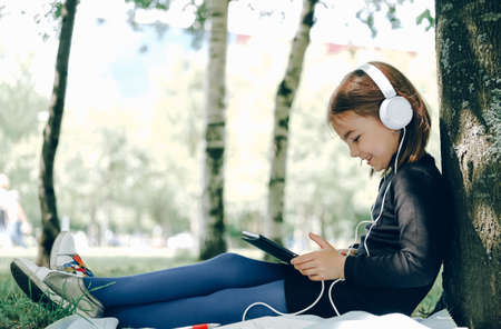 happy girl in white headphones using digital tablet pc in the park. Distant learning concept. Resilience, back to school, new normal.の写真素材