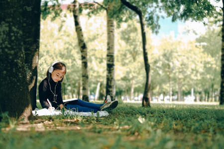 happy girl in white headphones using digital tablet pc in the park. Distant learning concept. Resilience, back to school, new normal.の写真素材