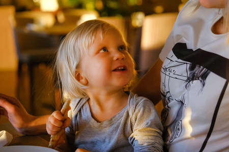 Young blond mother relaxing in sunny indoor cafe text messaging with her phone with her little cute child eating pizza.の写真素材