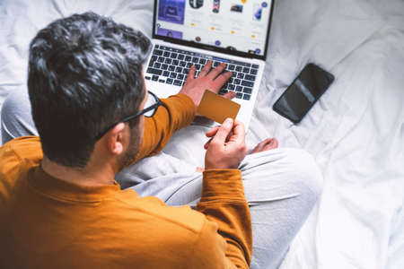 Man in yellow pajamas sitting at the table with credit card and smart phone shopping online and payingの写真素材