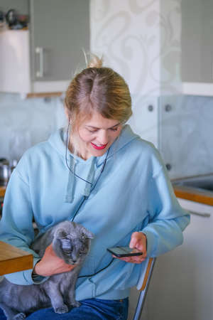 Young business woman in blue sweatshort with gray cat looking at cell phone working at home with computer in the kitchenの写真素材