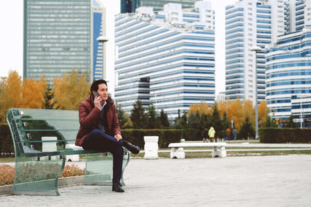 Middle aged asian man wearing autumn clothes sitting on the bench with his phone in downtown.の写真素材