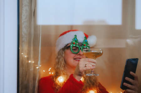 Happy woman in Santa hat and masquerade glasses with a glass of champane celebrating New Year's Eve or Christmas making video callの写真素材