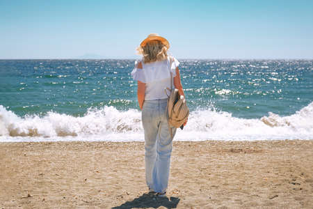 Woman in straw hat and blue jeans walking along seaside.