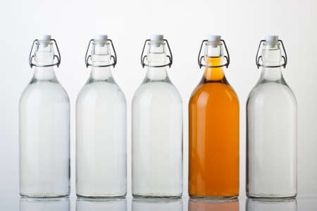 Five glass bottles on white background. Four with clear water, one with orange colored liquid. の写真素材