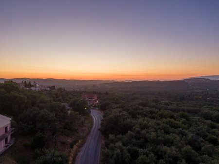 Aerial view of a winding road through a forest at sunset, with a village and mountains in the distance.の写真素材
