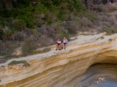 Two people sit on a cliff edge overlooking a rocky cove, surrounded by lush greenery.の写真素材