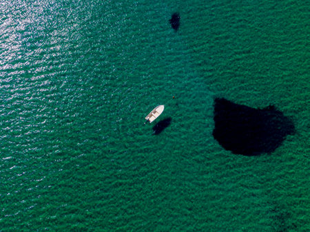 Aerial view of a small boat on a calm, teal sea. Dark underwater formations are visible.