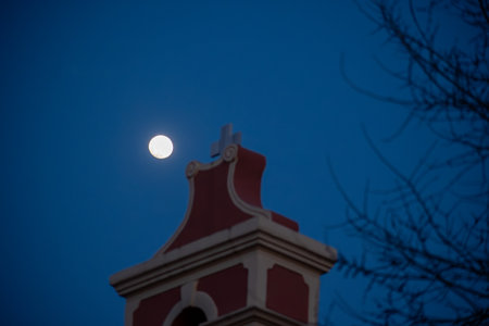 Moon glows beside building with cross, deep blue sky providing backdrop with tree silhouette.の写真素材