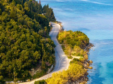 Coastal road winds beside turquoise waters, embraced by lush green trees and rocky shoreline. Serene.の写真素材