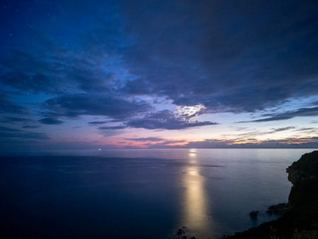 Moonlight pierces through a dramatic, cloud-filled sky, illuminating the dark ocean below at dusk.