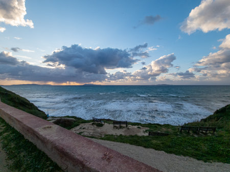 Coastal view featuring dramatic skies over the ocean, benches line the cliffside viewpoint area.の写真素材