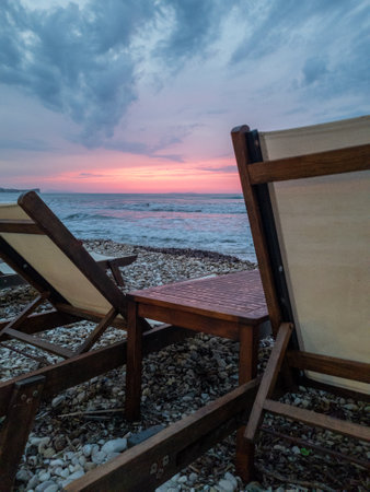 Relaxing view of ocean waves meeting pebble beach, lounge chairs, and sunset sky in backgroundの写真素材