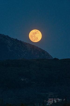 Full moon rising above a dark, forested mountain range illuminating the dark, night sky.の写真素材