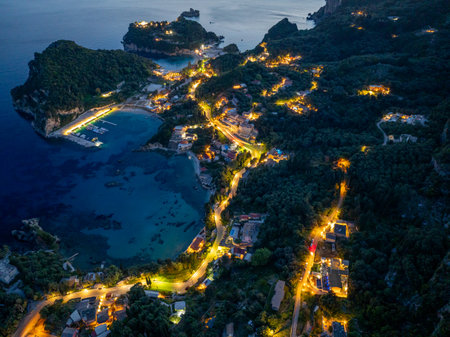 Night aerial view of a coastline village featuring illuminated buildings, roads, and a tranquil harbor.の写真素材