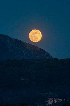 A luminous moon rises over a dark, shadowed mountain range at dusk, casting a golden glow over the scene.の写真素材