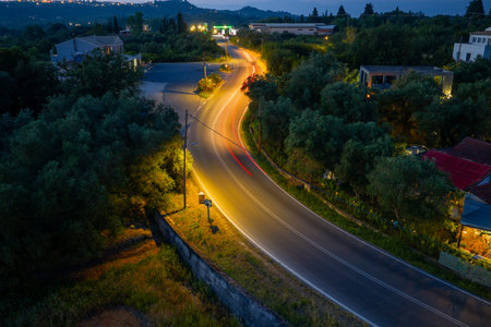 A curving asphalt road illuminated by streetlights at dusk, with blurred lights indicating passing vehicles.の写真素材