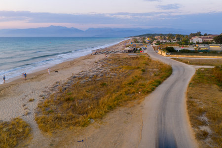 Serene Ionian Coastline at Sunset: An aerial view capturing the tranquil beauty of a Greek beach with gentle waves, distant mountains, and a peaceful road as dusk settles.の写真素材