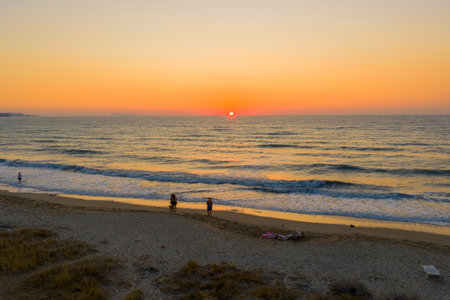 Vibrant Golden Hour on the Ionian Coast: A captivating beach scene with a fiery sunset over the sea, people relaxing by the shore, and gentle waves lapping the sand.の写真素材