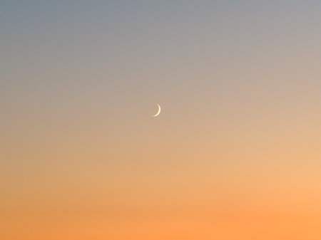 Crescent Moon in the Evening Sky: A serene and minimalist shot of a thin, glowing crescent moon set against a soft, gradient sky during twilight, capturing the peaceful transition from day to night.の写真素材