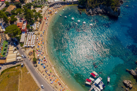 High angle aerial shot of a busy beach in Paleokastritsa, Corfu. Sun glitters on the clear turquoise sea as people swim, sunbathe under umbrellas, and boats wait by the shore. Perfect summer sceneの写真素材