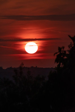 A moody, deep red sunset in a vertical composition. The bright sun hangs in a dark, fiery sky above a black silhouette of hills and foliage, creating a dramatic and intense end-of-day sceneの写真素材