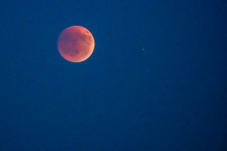 Stunning view of the Blood Moon during a total lunar eclipse, glowing a deep red-orange against the dark blue night sky. A rare and beautiful celestial event with faint stars visibleの写真素材