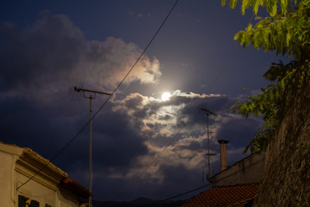 A dramatic night in a Greek village, with the full moon illuminating a chaotic sky of dark clouds. Rooftops, antennas, and chimneys create a moody, urban silhouetteの写真素材