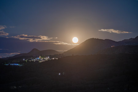 A magical full moon rises over a serene Greek village nestled in the mountains. The bright moonlight illuminates the landscape under a starry night sky, creating a tranquil and enchanting scene.の写真素材