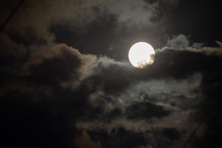 The brilliant full moon shines like the sun through a break in dark, moody clouds, creating a...