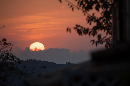A tranquil sunset viewed from a balcony or window, framed by dark silhouettes of leaves. The golden sun sinks behind clouds and distant hills with a castle in a serene, picturesque landscapeの写真素材