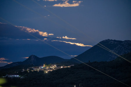 A tranquil Greek village glows in a mountain valley at night, under a starry sky with softly lit clouds. Strange light streaks add a unique, futuristic effect to this serene landscapeの写真素材