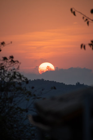 A picturesque sunset over the Italian countryside, with the large orange sun framed by soft foliage. A distant historic villa sits atop the silhouetted hills of Tuscany, creating a romantic sceneの写真素材