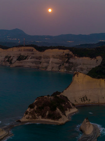 Vertical aerial shot of a bright full moon rising over the dramatic white cliffs of Cape Drastis, Corfu. The sea stacks and coast are lit by the twilight skyの写真素材